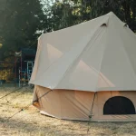 A large beige bell tent set up in a grassy open area with a nearby vehicle and rooftop tent under shaded trees.