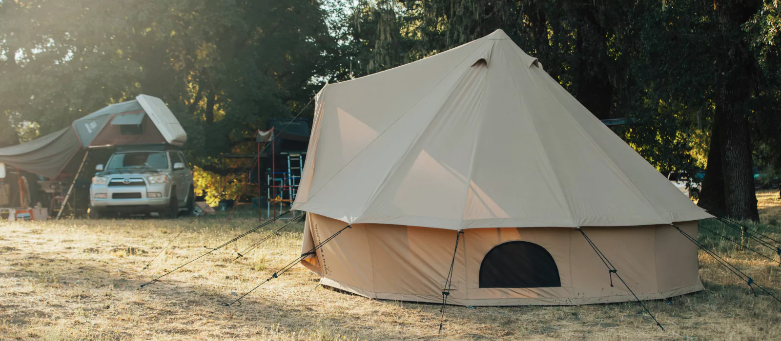 A large beige bell tent set up in a grassy open area with a nearby vehicle and rooftop tent under shaded trees.