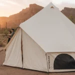 Guests enjoying a birthday camping party in the desert with a large canvas tent and mountain views at sunset.