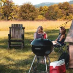 Friends relaxing near a bell tent with a grill and cooler set up for camp cooking in a sunny open field.