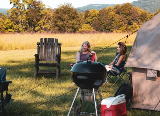Friends relaxing near a bell tent with a grill and cooler set up for camp cooking in a sunny open field.