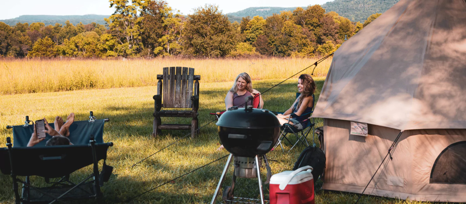 Friends relaxing near a bell tent with a grill and cooler set up for camp cooking in a sunny open field.