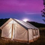 A glowing wall tent set up under a starry night sky, surrounded by trees and mountains, perfect for camping in Florida.