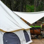 Camper preparing a meal at a campsite with a bell tent and tarp shelter in a forest setting.