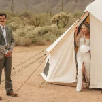 Bride stepping out of a canvas tent in a desert setting while the groom stands nearby smiling.