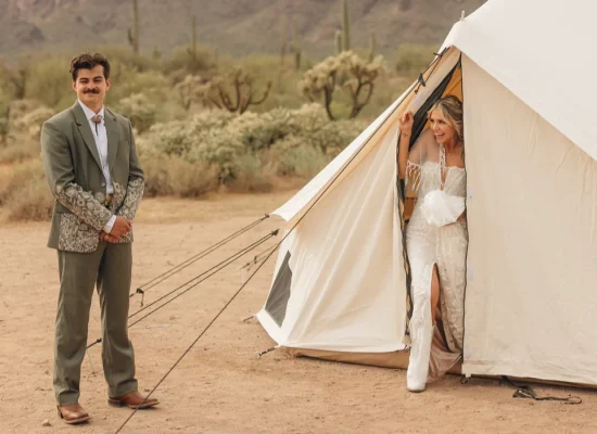 Bride stepping out of a canvas tent in a desert setting while the groom stands nearby smiling.
