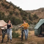 Two friends walking their bikes near tents at a scenic campsite surrounded by mountains and desert vegetation.