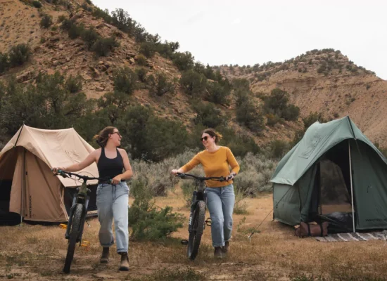 Two friends walking their bikes near tents at a scenic campsite surrounded by mountains and desert vegetation.