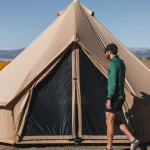 A man in outdoor clothing walks toward a large canvas bell tent set up in an open field with mountains and autumn trees in the background.