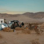 Group camping in Oregon desert with canvas tent, campfire, and vehicles under mountain sunset.