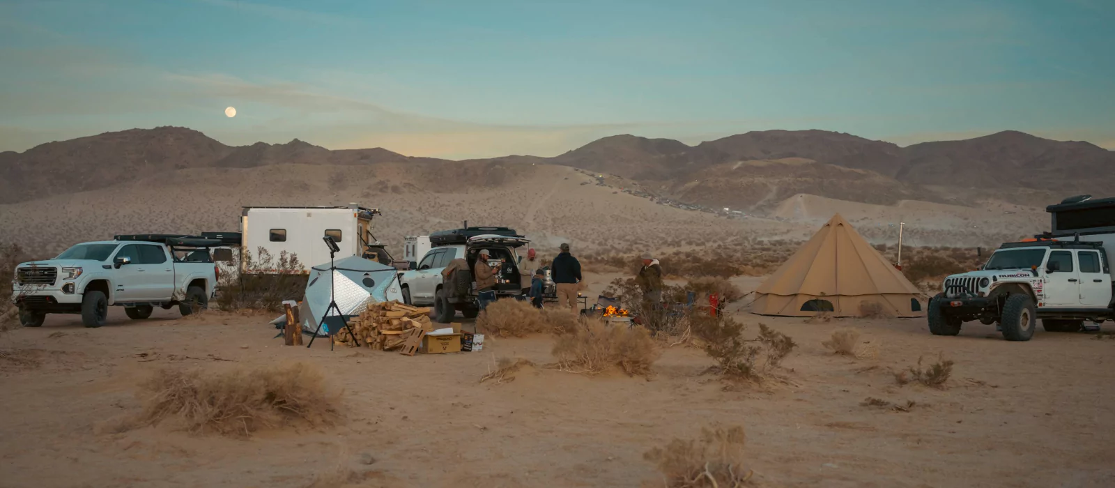 Group camping in Oregon desert with canvas tent, campfire, and vehicles under mountain sunset.