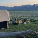 Family gathered around a campfire sharing stories on a fall camping night beside a canvas tent in the mountains.