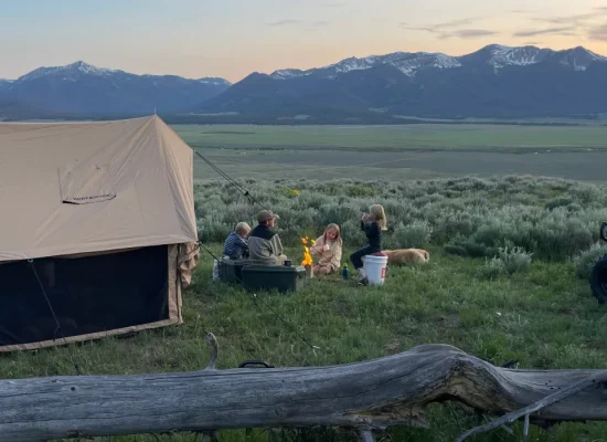 Family gathered around a campfire sharing stories on a fall camping night beside a canvas tent in the mountains.