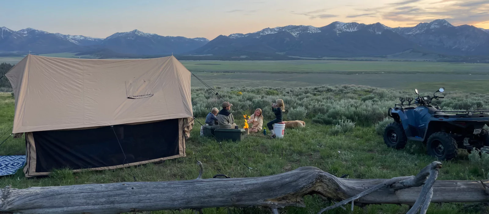 Family gathered around a campfire sharing stories on a fall camping night beside a canvas tent in the mountains.