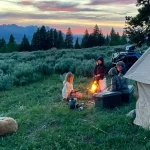 Family enjoying fall camping around a campfire with a canvas tent and dog at sunset in the mountains.