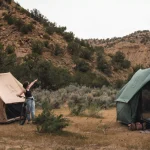 Two people set up canvas tents in a desert canyon during a fall camping trip, surrounded by rugged hills, dry brush, and mountain bikes.