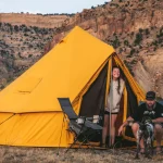A bright yellow canvas tent set up in a desert canyon landscape with campers and a dog enjoying the outdoors.