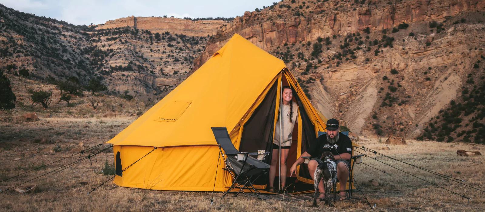 A bright yellow canvas tent set up in a desert canyon landscape with campers and a dog enjoying the outdoors.