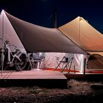 Bell tent illuminated at night with awning, camping chairs, and bike setup on a wooden platform in the wilderness.