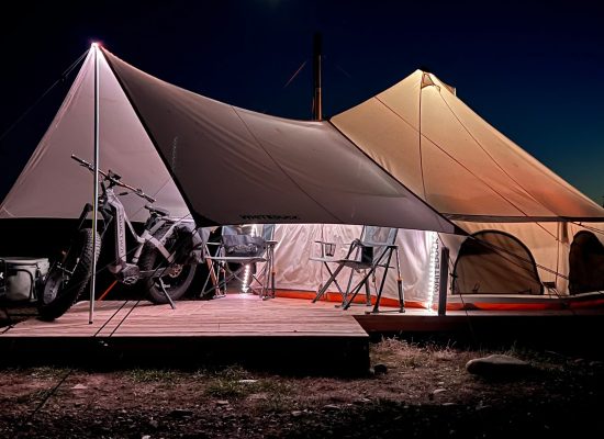 Bell tent illuminated at night with awning, camping chairs, and bike setup on a wooden platform in the wilderness.