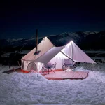 Cozy bell tent with awning set up for winter camping at night, surrounded by snow with mountains in the background.