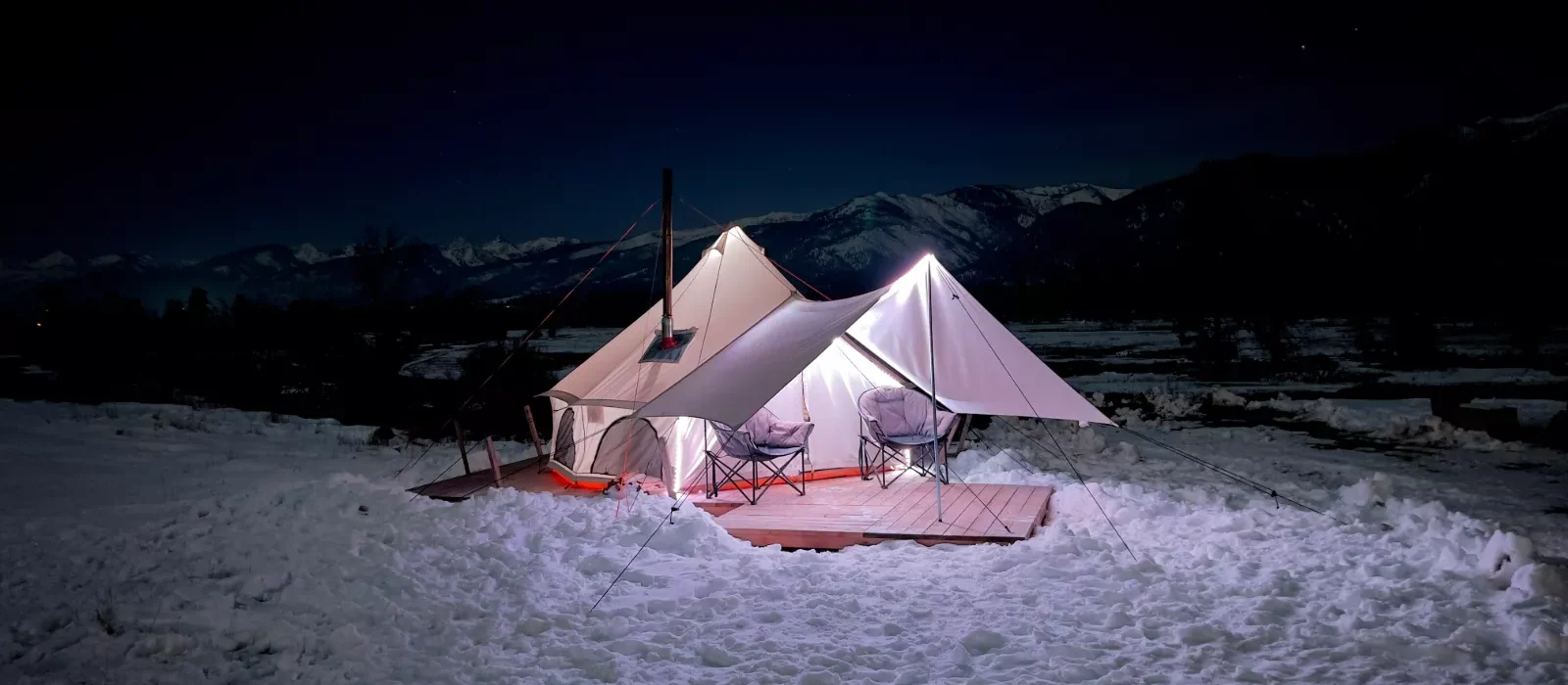 Cozy bell tent with awning set up for winter camping at night, surrounded by snow with mountains in the background.