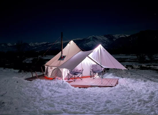 Cozy bell tent with awning set up for winter camping at night, surrounded by snow with mountains in the background.
