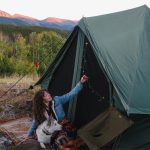 Woman camping with dog beside a canvas tent in the mountains at sunset with forest scenery