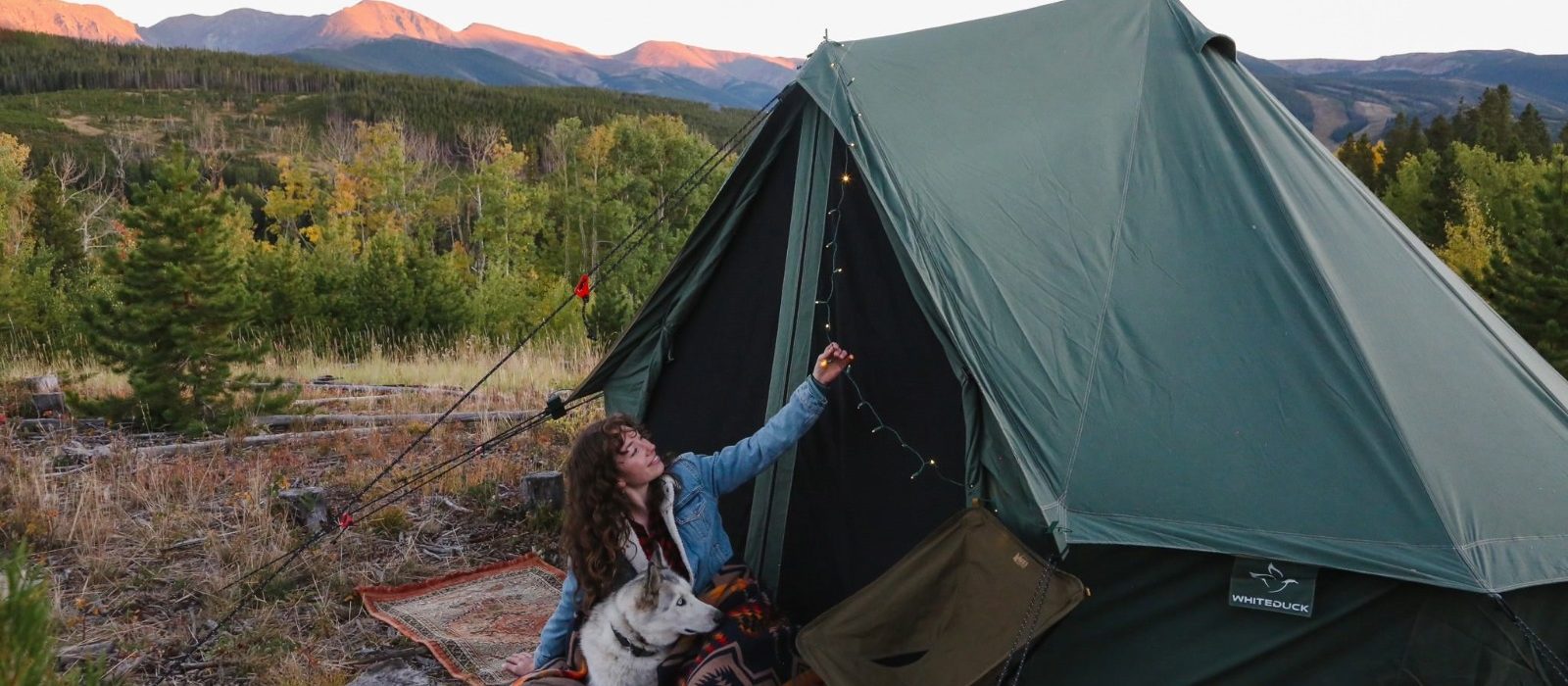 Woman camping with dog beside a canvas tent in the mountains at sunset with forest scenery