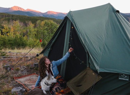 Woman camping with dog beside a canvas tent in the mountains at sunset with forest scenery