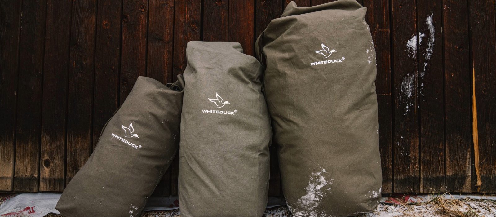 Three durable canvas camping bags from White Duck Outdoors resting against a wooden wall, designed for heavy-duty outdoor gear storage.