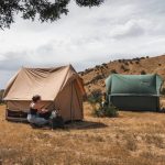 Person setting up a canvas scout tent at a scenic outdoor campsite with another tent in the background.