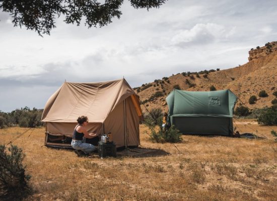 Person setting up a canvas scout tent at a scenic outdoor campsite with another tent in the background.