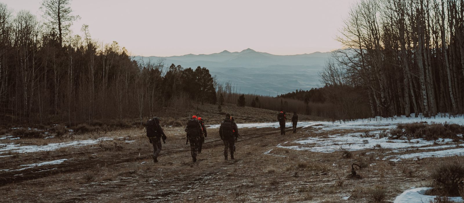 A group of people hiking together through a snowy field with trees and mountains in the distance, exploring the outdoors at sunset.