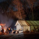A family sets up camp at night with a glowing campfire, wall tents, and tall trees under a starry sky, creating a cozy campsite base.