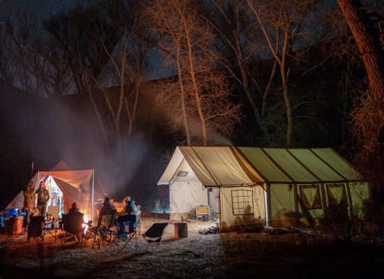 A family sets up camp at night with a glowing campfire, wall tents, and tall trees under a starry sky, creating a cozy campsite base.