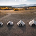 High-angle drone shot of four canvas bell tents of varying sizes lined up in a dry, grassy field, with a forest of yellow and orange autumn trees and distant mountains under a blue sky in the background.