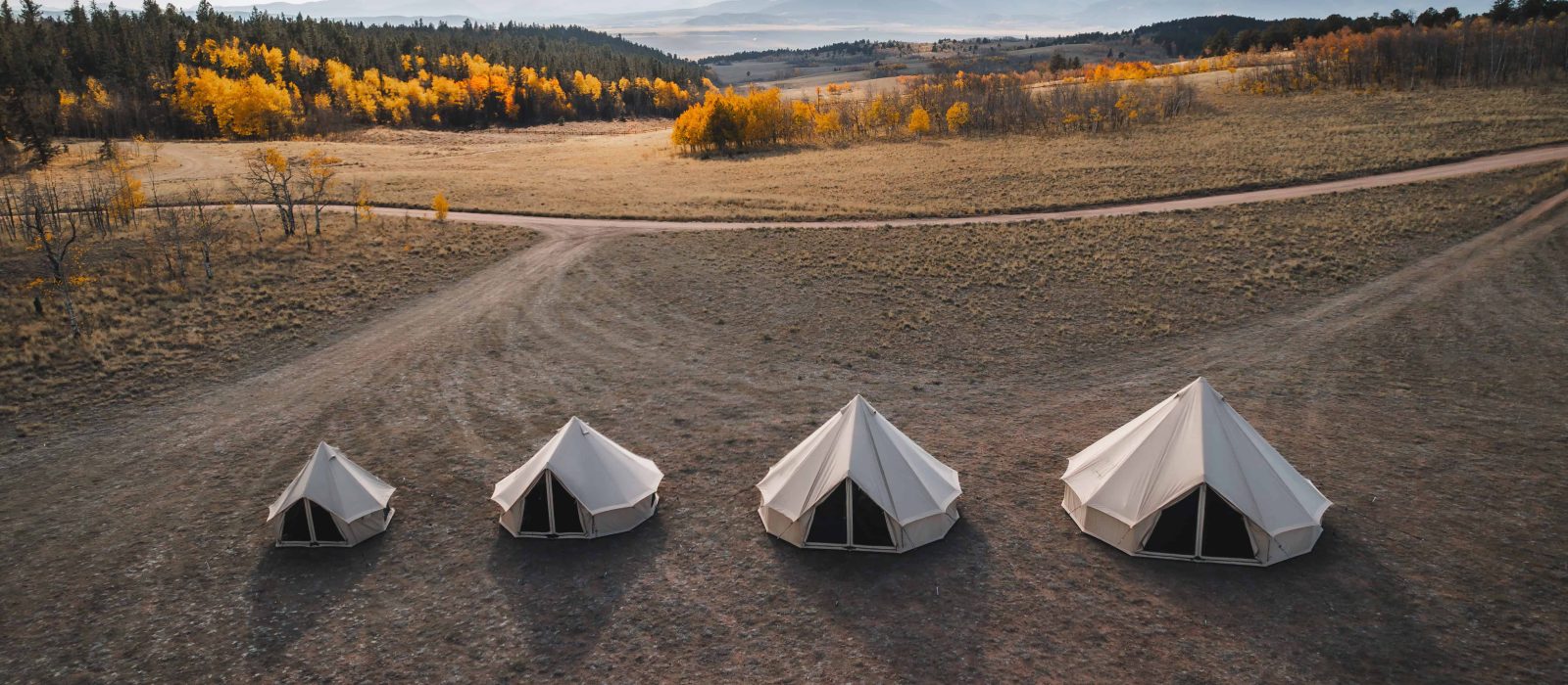 High-angle drone shot of four canvas bell tents of varying sizes lined up in a dry, grassy field, with a forest of yellow and orange autumn trees and distant mountains under a blue sky in the background.