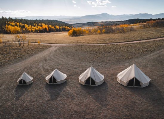 High-angle drone shot of four canvas bell tents of varying sizes lined up in a dry, grassy field, with a forest of yellow and orange autumn trees and distant mountains under a blue sky in the background.