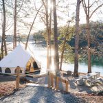 Glamping bell tent set up on a wooden platform beside a sunny lake, surrounded by a sparse forest with a picnic table and chairs in the foreground.