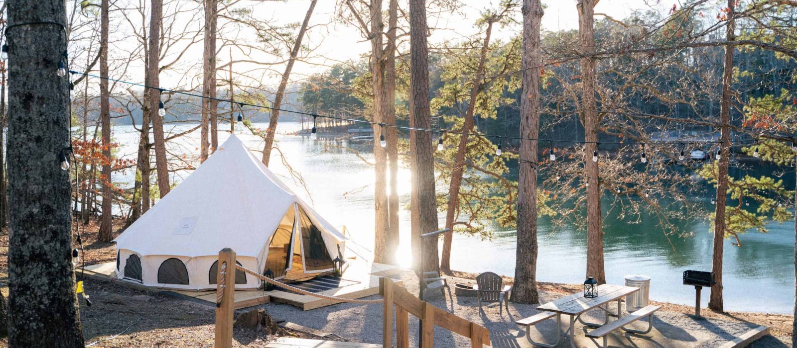 Glamping bell tent set up on a wooden platform beside a sunny lake, surrounded by a sparse forest with a picnic table and chairs in the foreground.