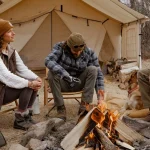 Campers relaxing around a campfire in front of a canvas tent with a log carrier filled with firewood beside them.