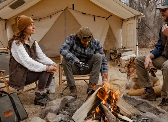 Campers relaxing around a campfire in front of a canvas tent with a log carrier filled with firewood beside them.