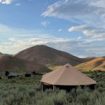 A military surplus tent set up in a scenic mountain valley surrounded by rolling hills under a bright blue sky.