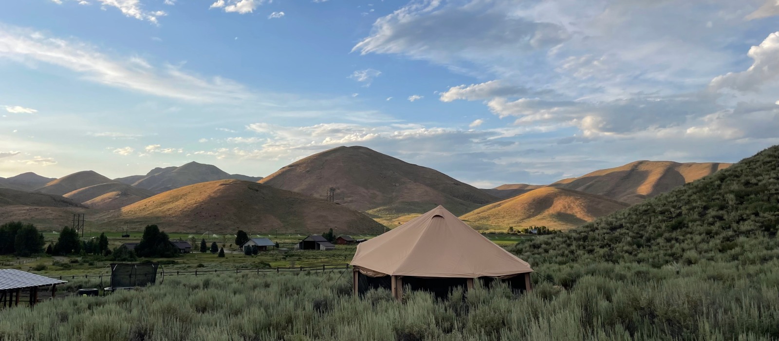 A military surplus tent set up in a scenic mountain valley surrounded by rolling hills under a bright blue sky.