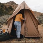 Person entering a scout tent set up in a dry, grassy outdoor area with camping gear nearby