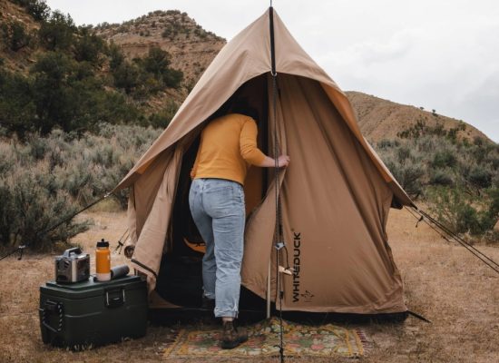 Person entering a scout tent set up in a dry, grassy outdoor area with camping gear nearby