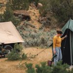 Camper setting up a green scout tent beside a tan canvas scout tent and mountain bikes in a scenic outdoor campsite.