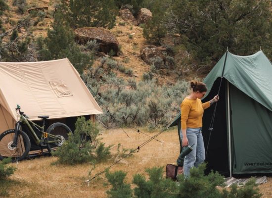 Camper setting up a green scout tent beside a tan canvas scout tent and mountain bikes in a scenic outdoor campsite.