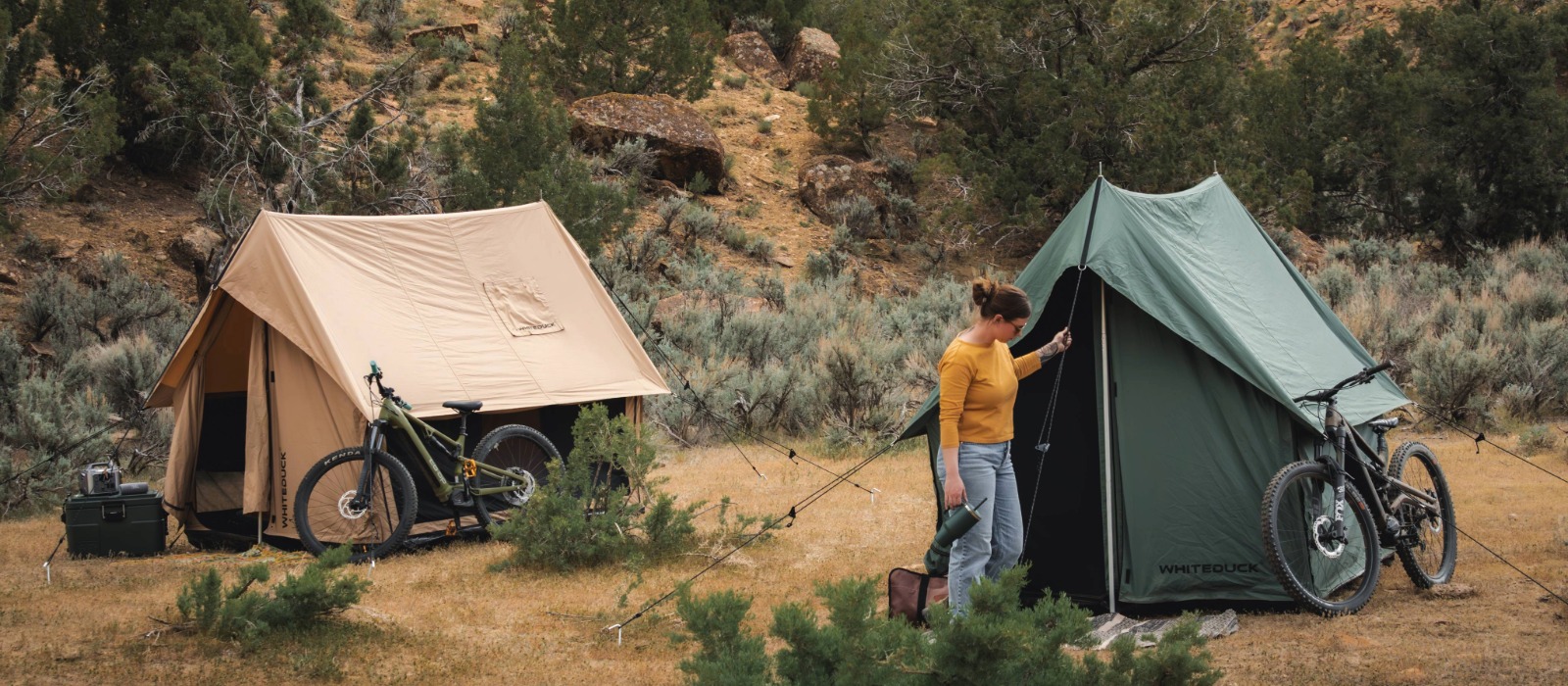 Camper setting up a green scout tent beside a tan canvas scout tent and mountain bikes in a scenic outdoor campsite.